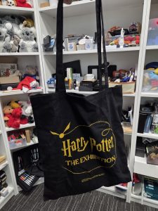 Black tote bag printed with a gold Harry Potter: The Exhibition design on the front.