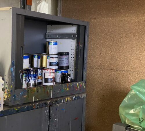 Open cabinet with paint cans and bottles, splattered counter, and print setup fees sign nearby.