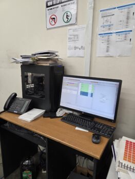 Office desk with a computer, phone, papers, and a note about Print Setup Fees on the wall.