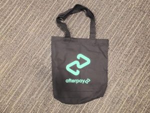 Black tote bag printed with the Afterpay logo in mint green on the front.