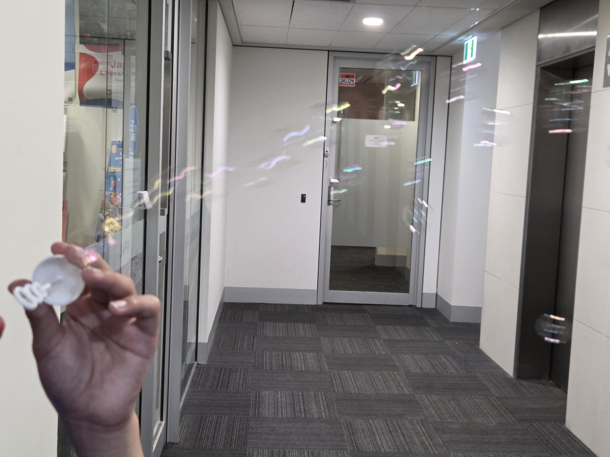 A bubble blower creates soap bubbles in a hallway near glass doors and office walls.