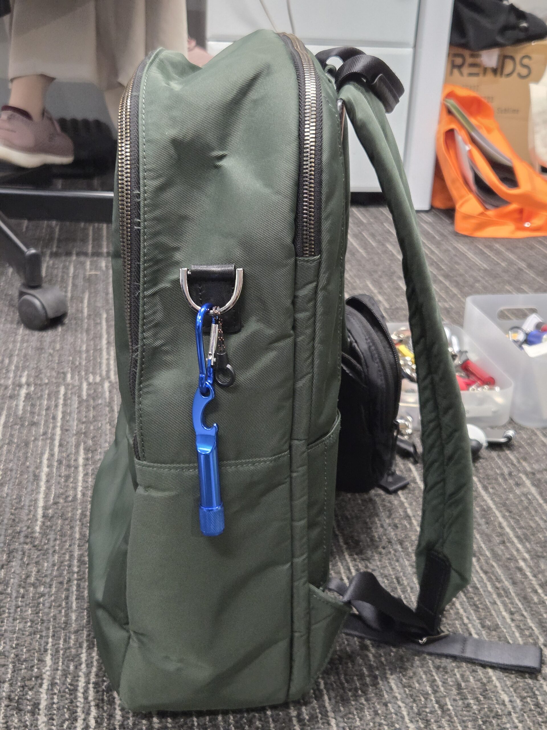 Green backpack with a blue carabiner and keyrings, sitting on an office floor near a desk.