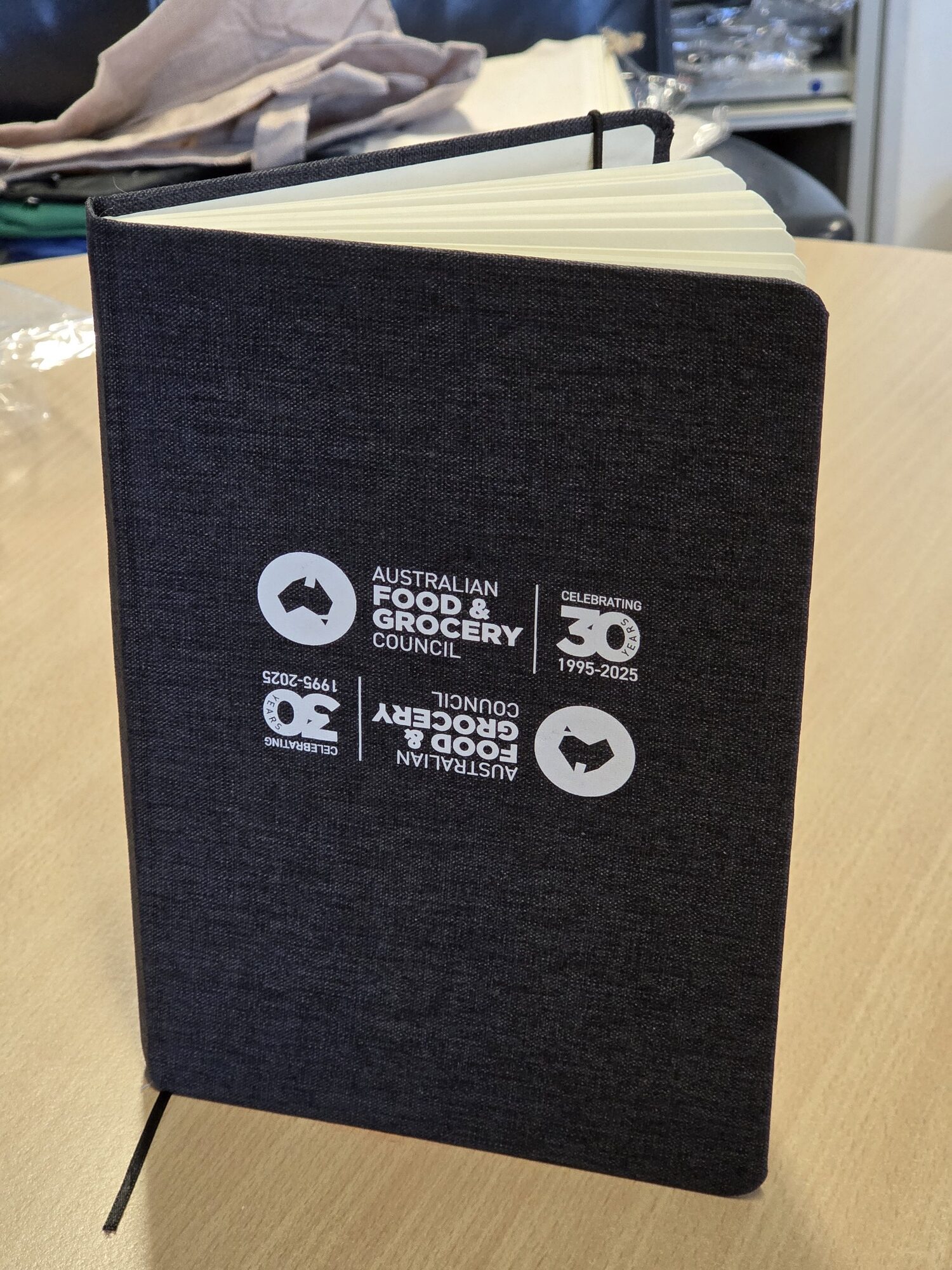 Black notebook with Australian Grocery Council logo and 1995-2025 printed on the cover.