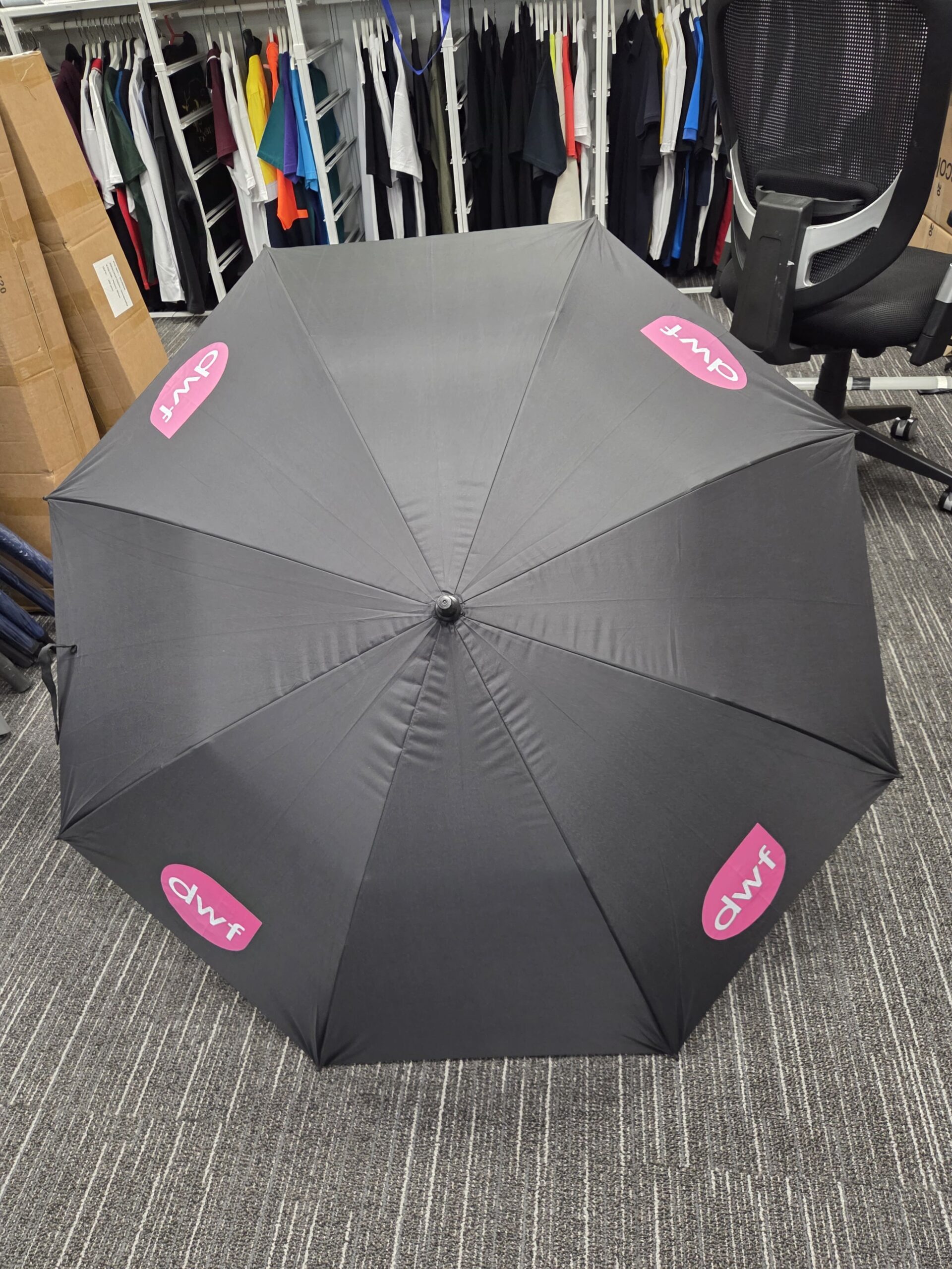 Black promotional products umbrella with pink dwp logos, open near a chair and clothes rack.