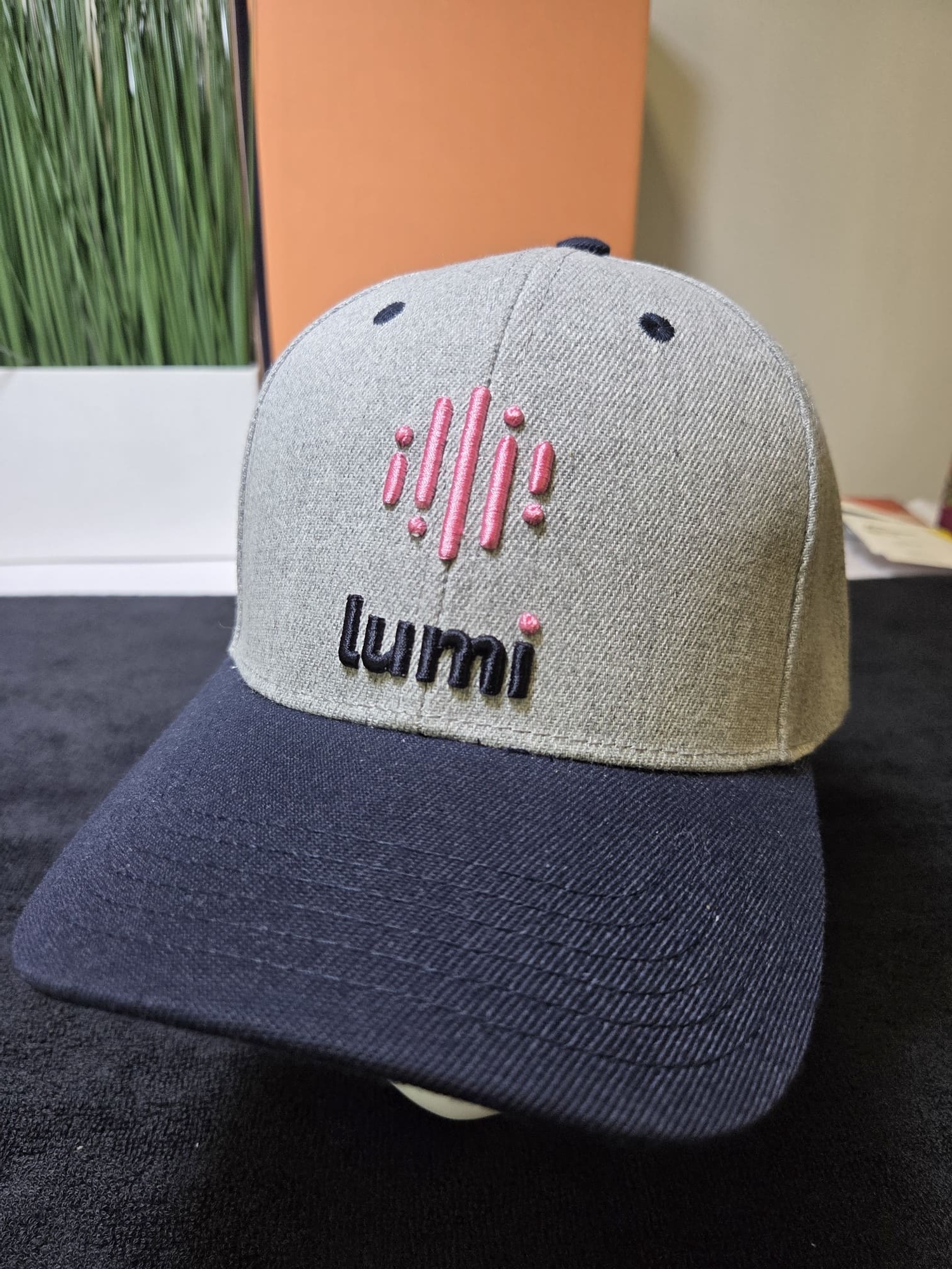 Gray and navy Lumi cap with pink and black logo on the front, placed on a dark surface.