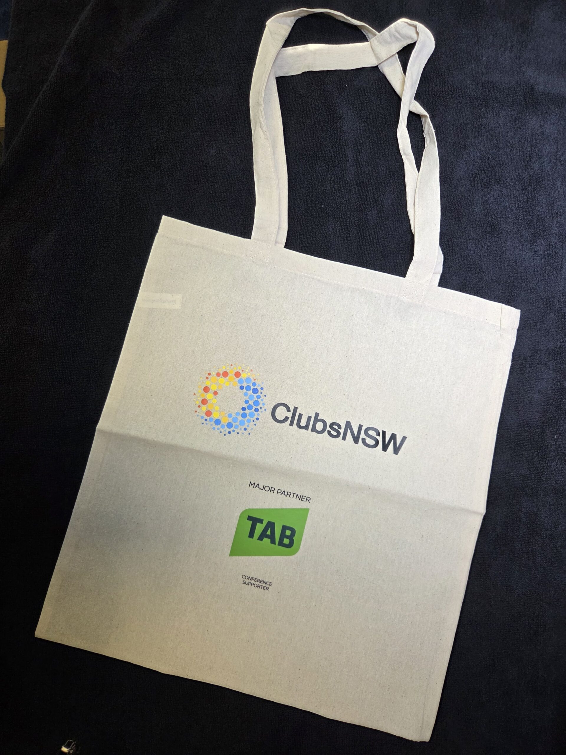 White tote bag featuring ClubsNSW and TAB logos on a dark fabric background.