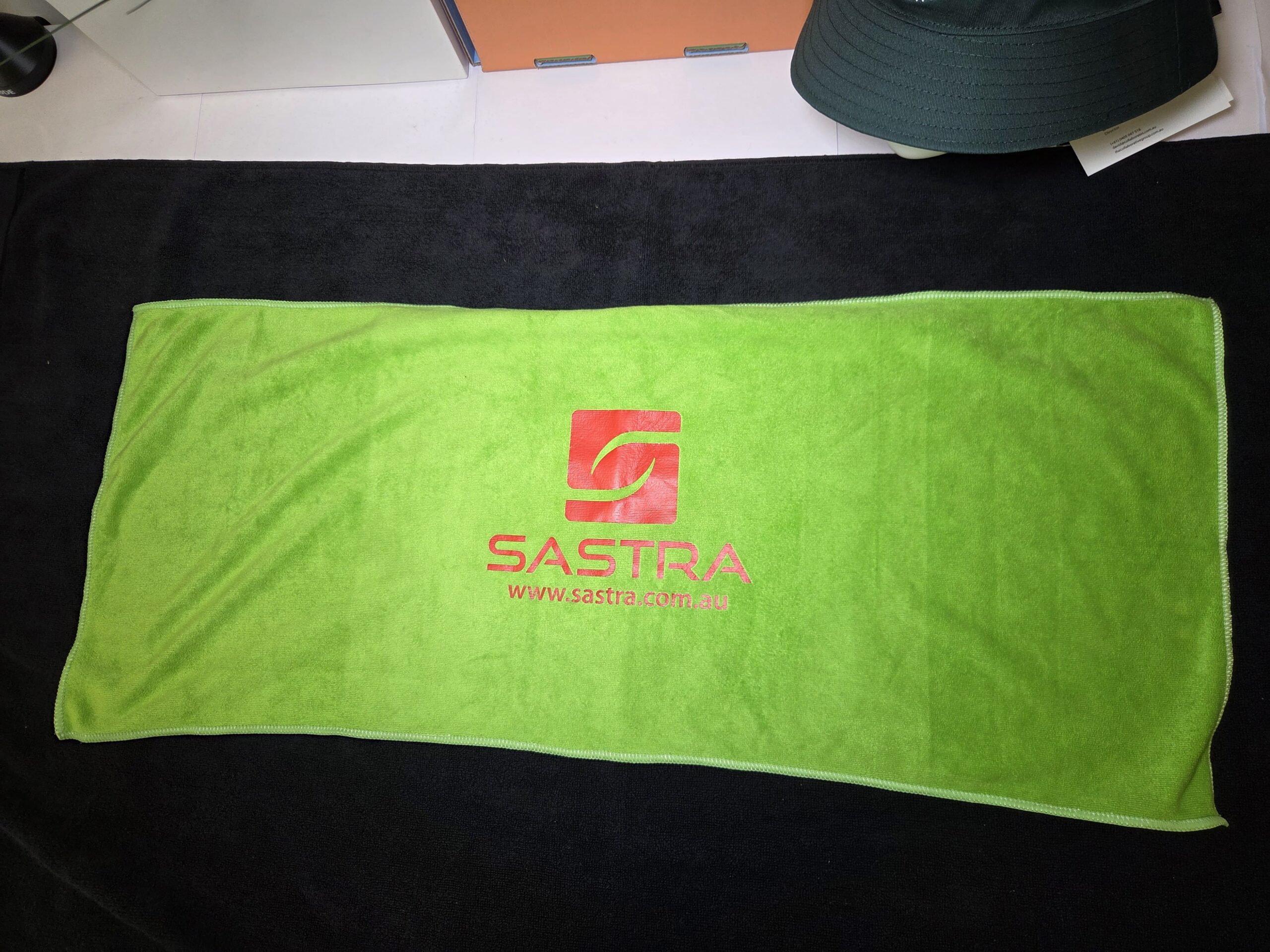 Bright green Sastra towel with logo and website in red, laid on a black surface.