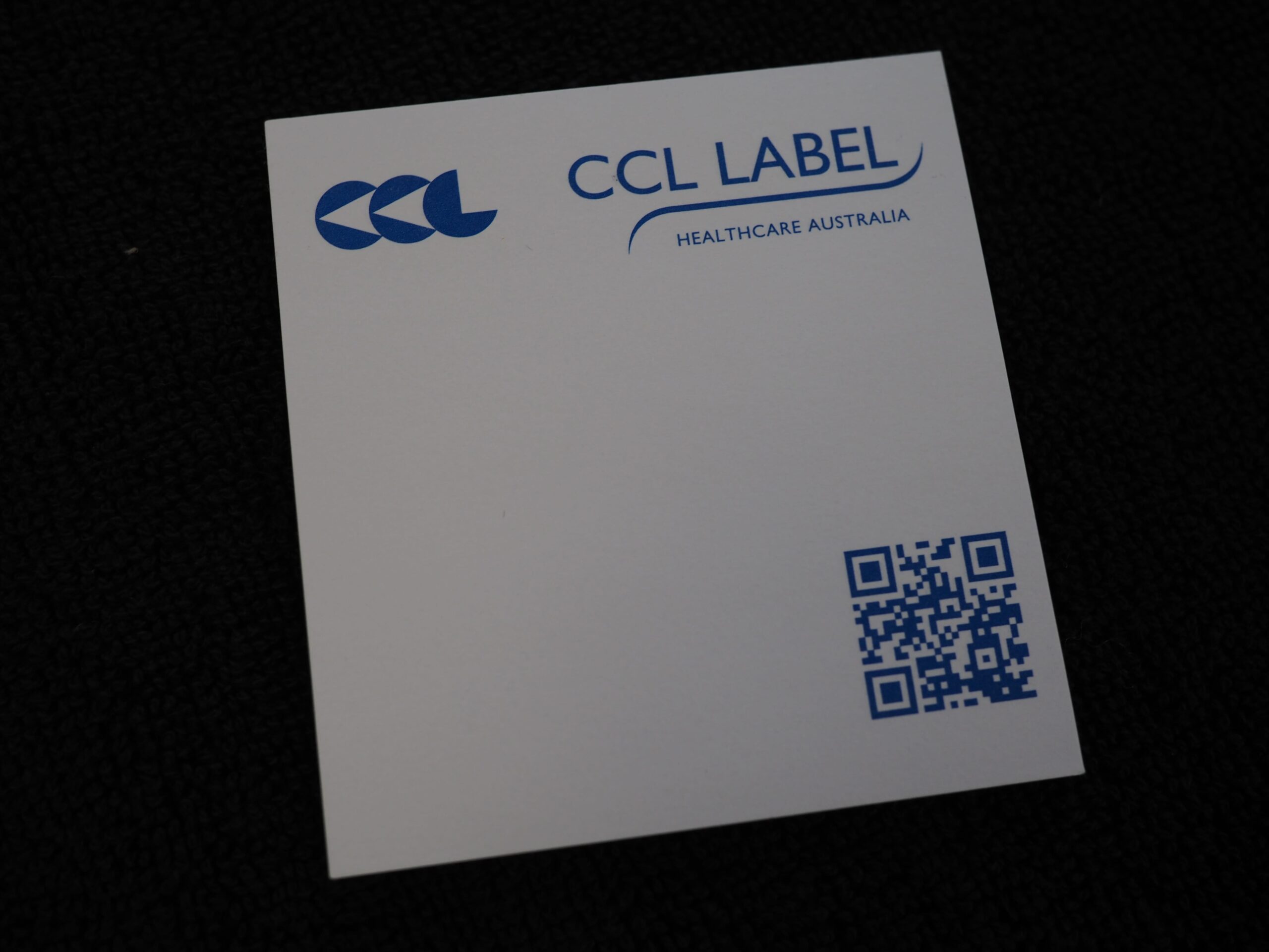 White notepad with CCL Label Healthcare branding and a blue QR code on a black surface.