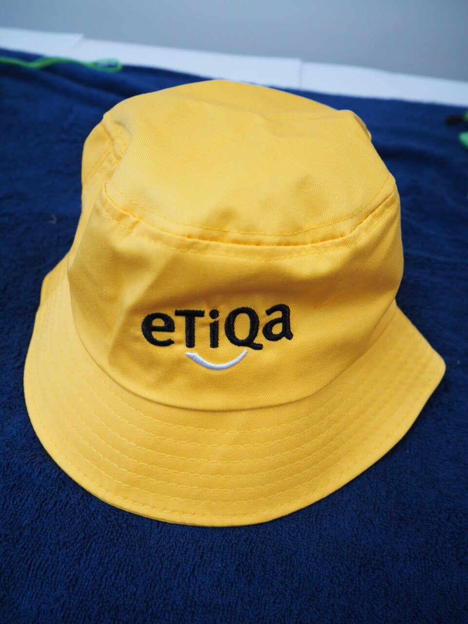 Yellow bucket hat with Etiqa Australia logo embroidered, placed on a dark blue towel.