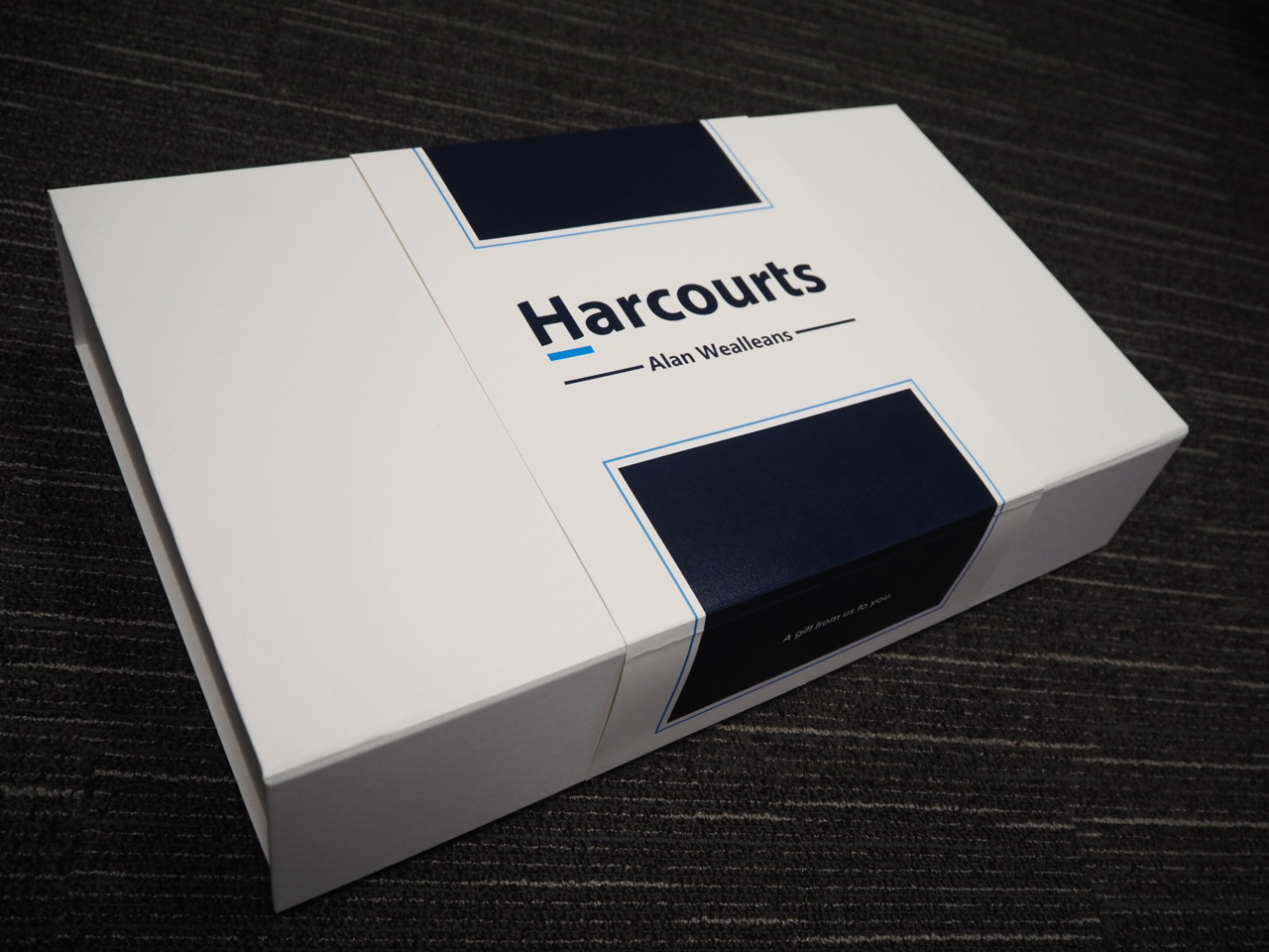 A white gift box with Harcourts Alan Wealleans branding on a dark carpeted floor.