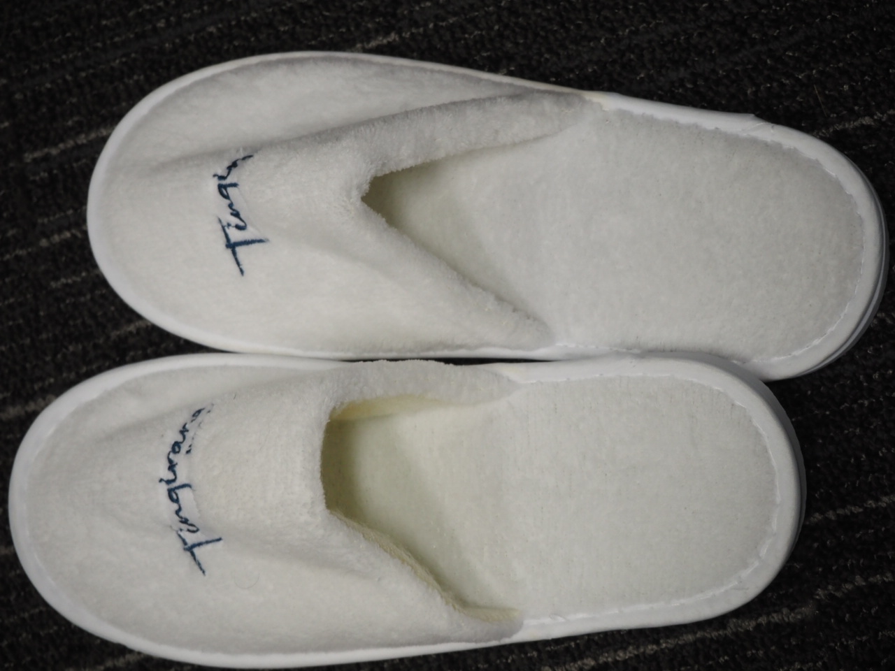 A pair of white hotel slippers with Tingirana Noosa blue writing, on a dark textured carpet.