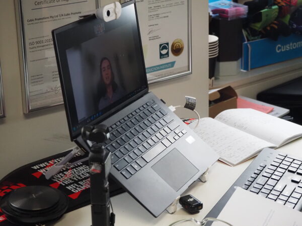 Laptop on a desk with a video call, notebooks, webcam, and promotional products nearby.