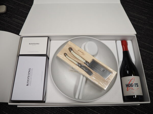 A corporate gift box with wine, plate, cheese knives, and two boxes on a dark striped surface.
