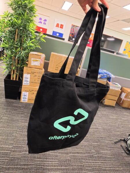 A hand holds a black Afterpay promotional products tote bag in an office with boxes and a plant.