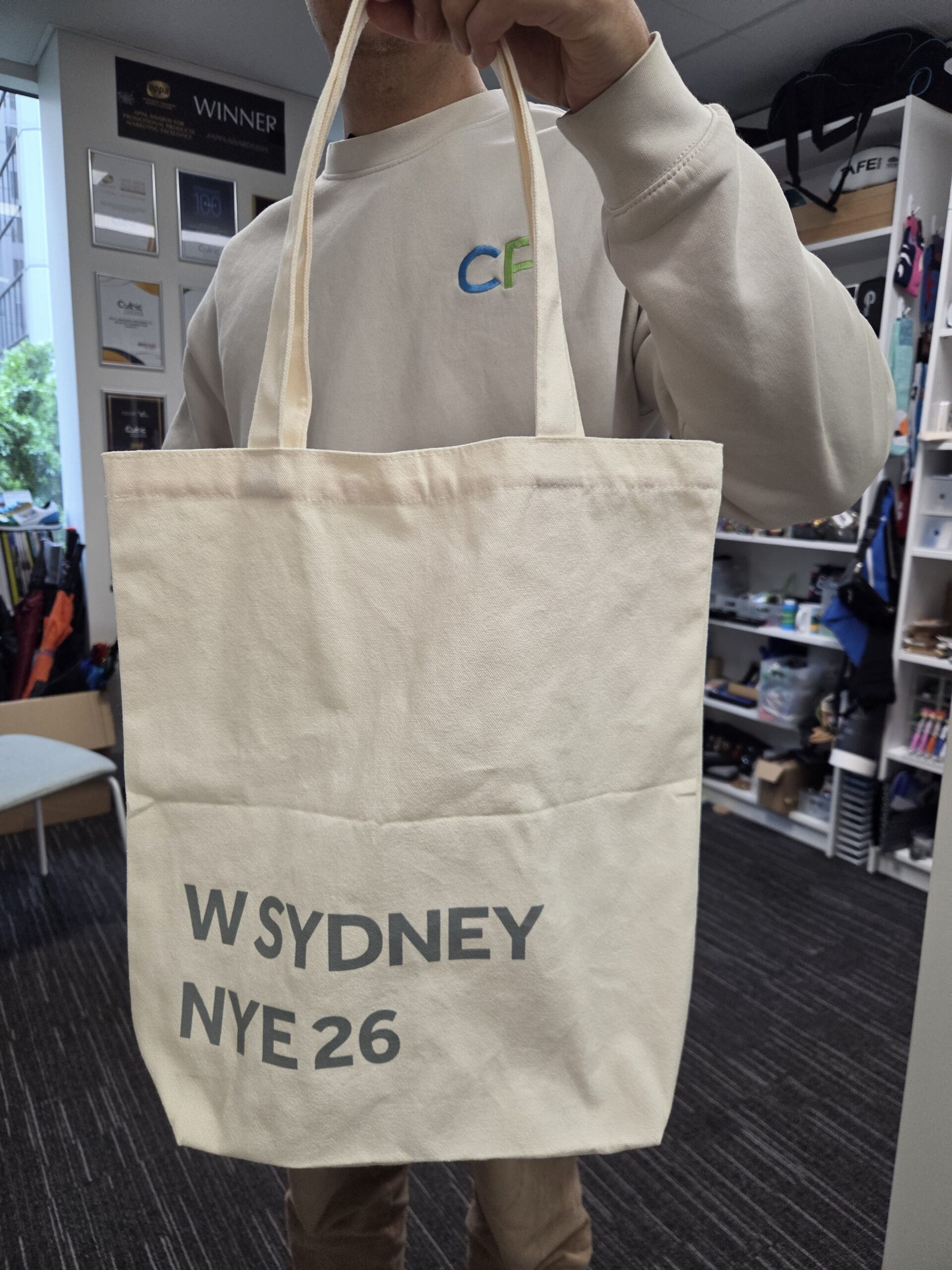Person holding a cream W Sydney tote bag labeled NYE 2026 in an office setting.