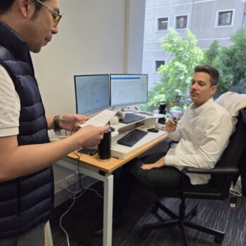 Two men discuss promotional merchandise in a modern office; one stands, the other sits at a desk.