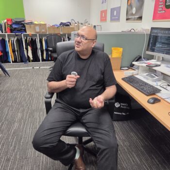 A man in black sits in an office chair, holding promotional merchandise near a desk with screens.