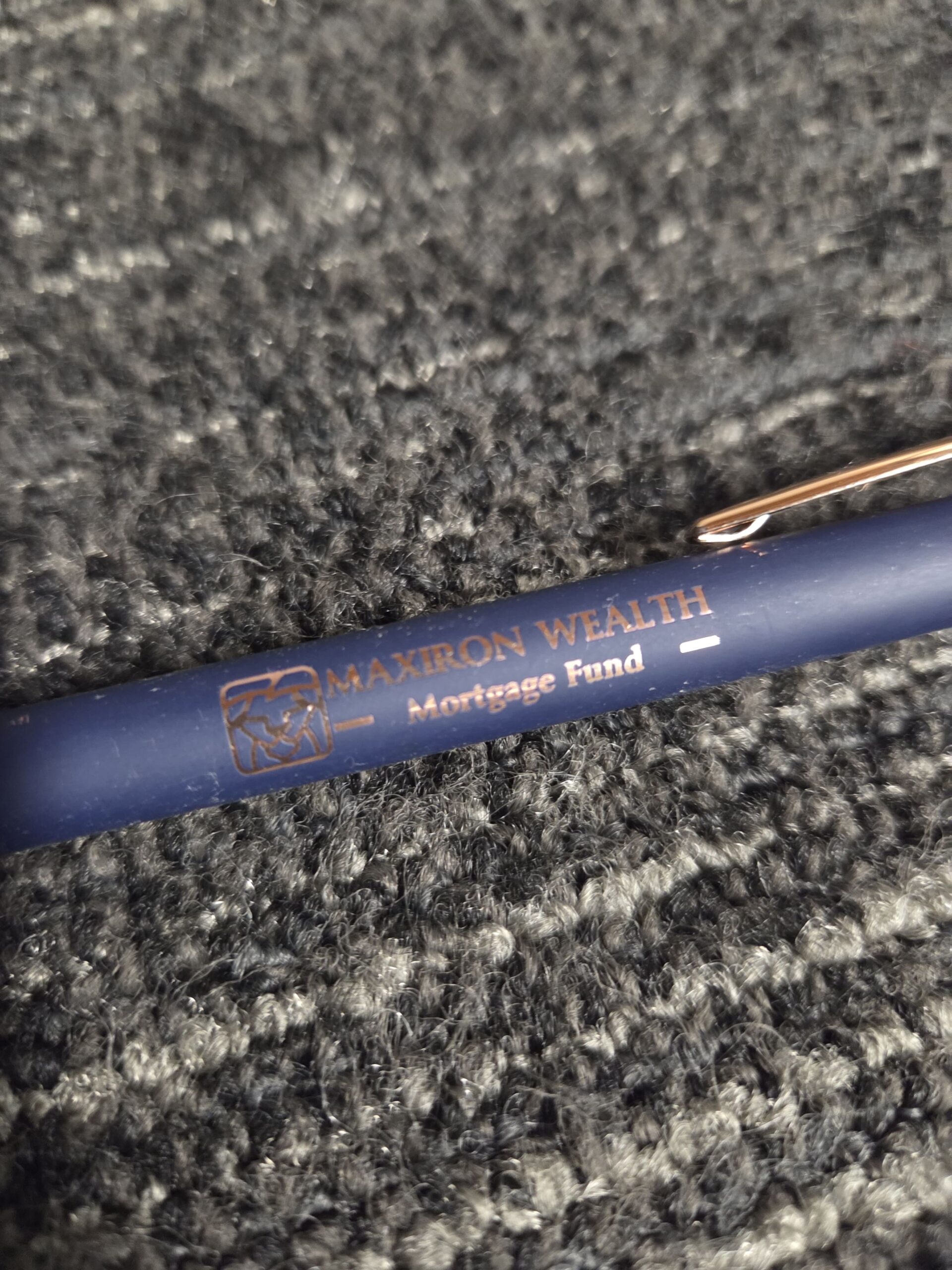 Blue pen on gray carpet with Maxiron Wealth branding in gold letters.