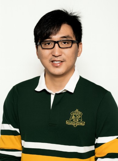 Man with glasses in a green and yellow collared shirt, standing against a white background.