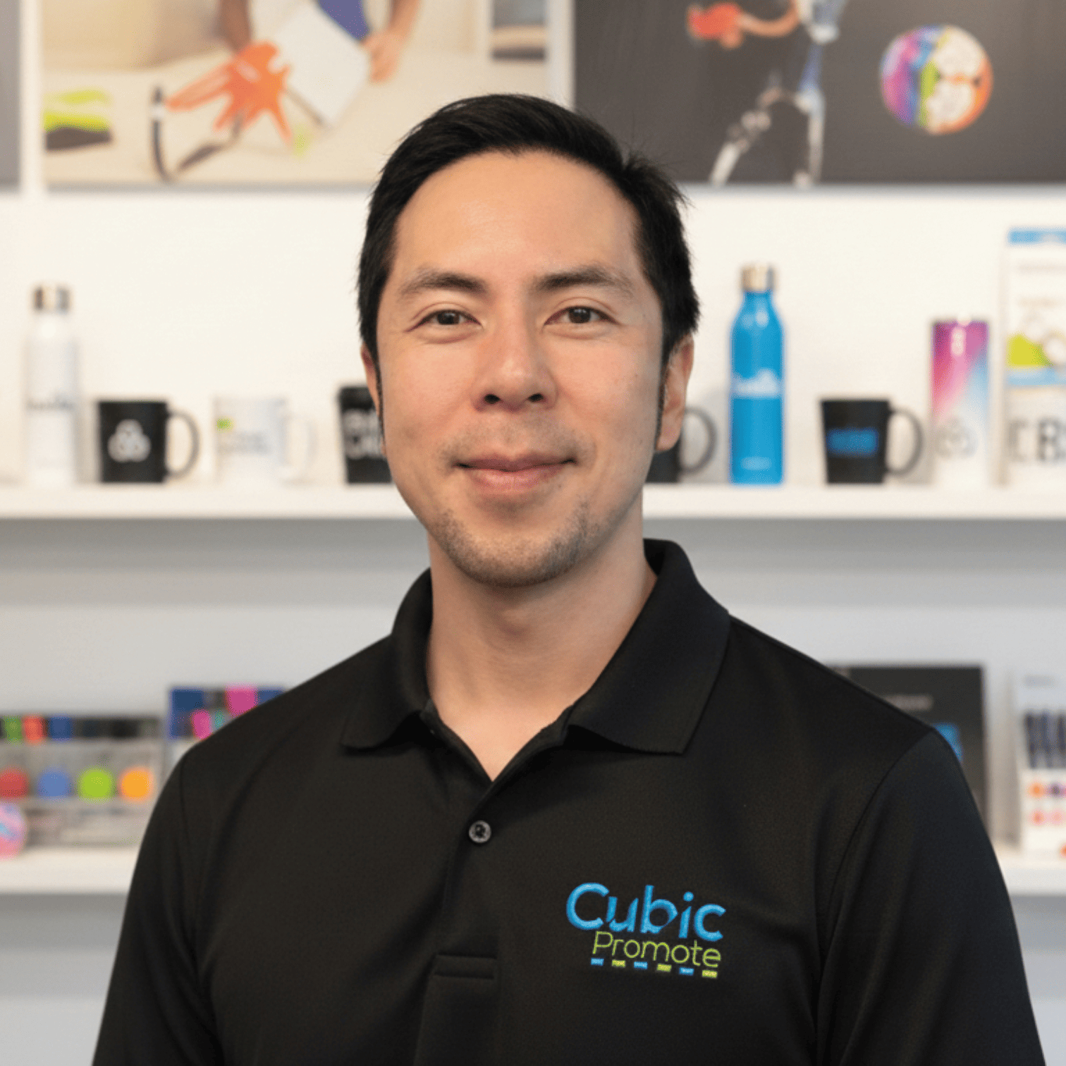 Meet the team: A man in a black Cubic Promote shirt smiles in an office with mugs and bottles behind him.