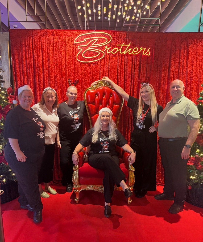Six people pose by a red throne at Brothers Leagues Club, under neon lights and Christmas trees.