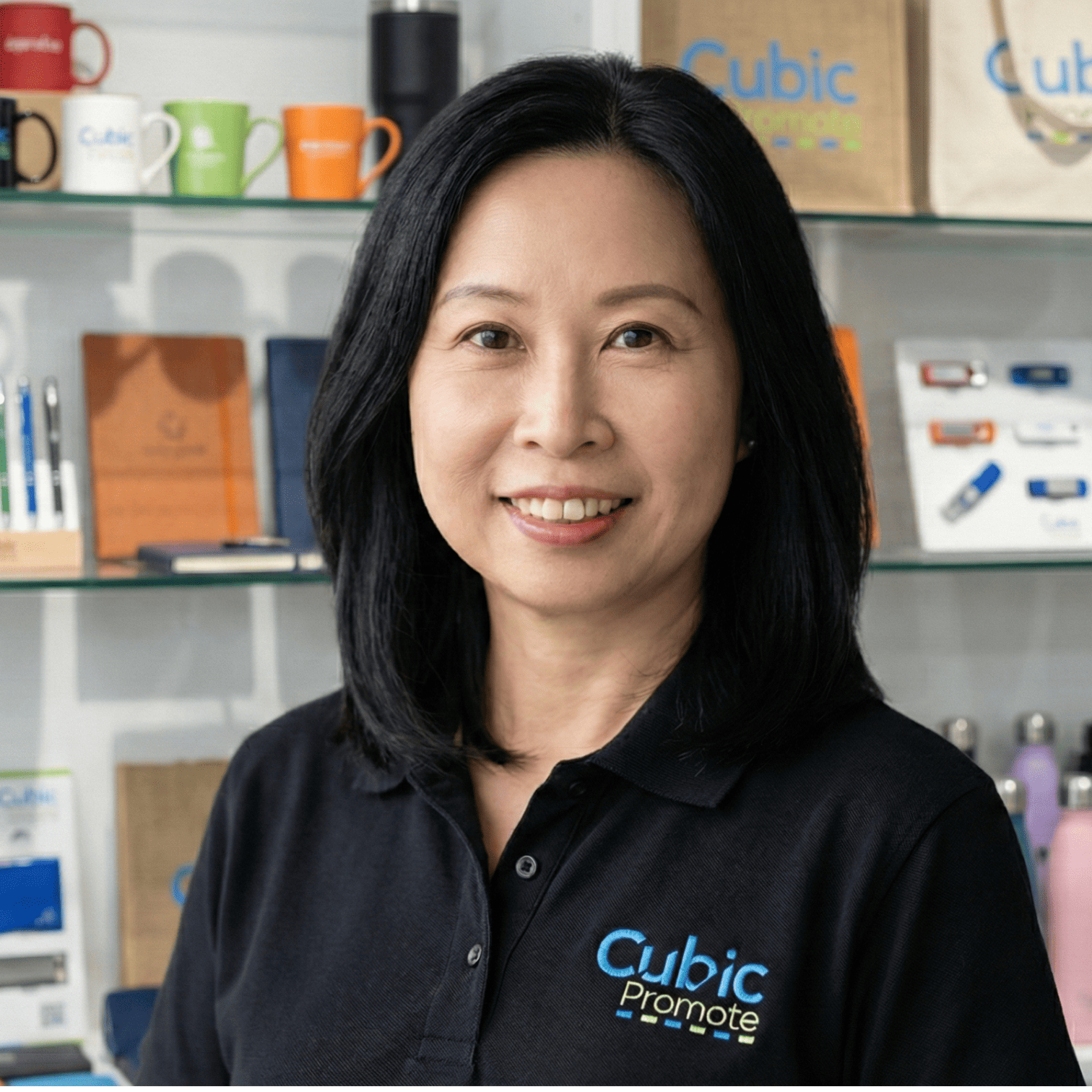 Smiling woman in a Cubic Promote shirt stands by branded merchandise shelves—meet the team!.