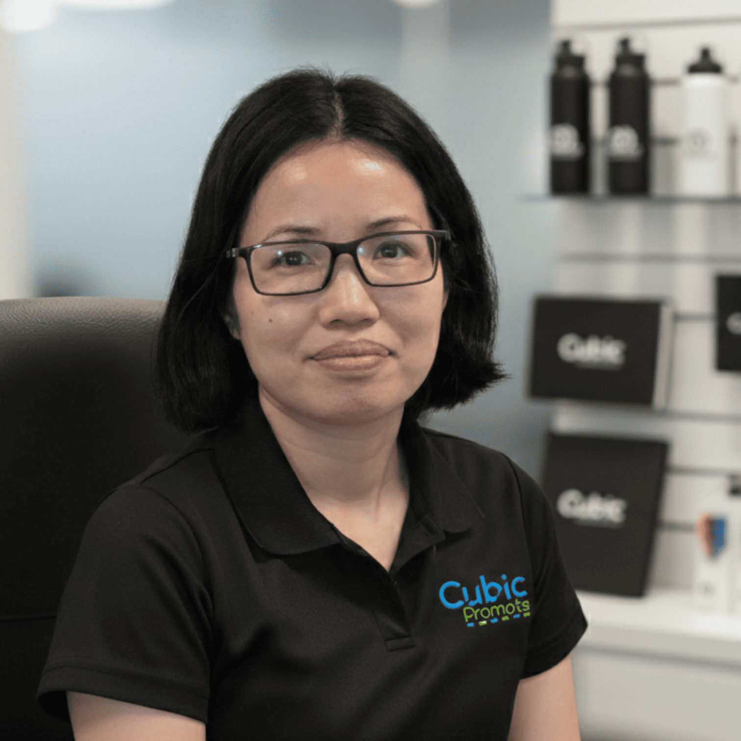 Meet the team: Woman with glasses in a black polo shirt sits in an office with branded items.