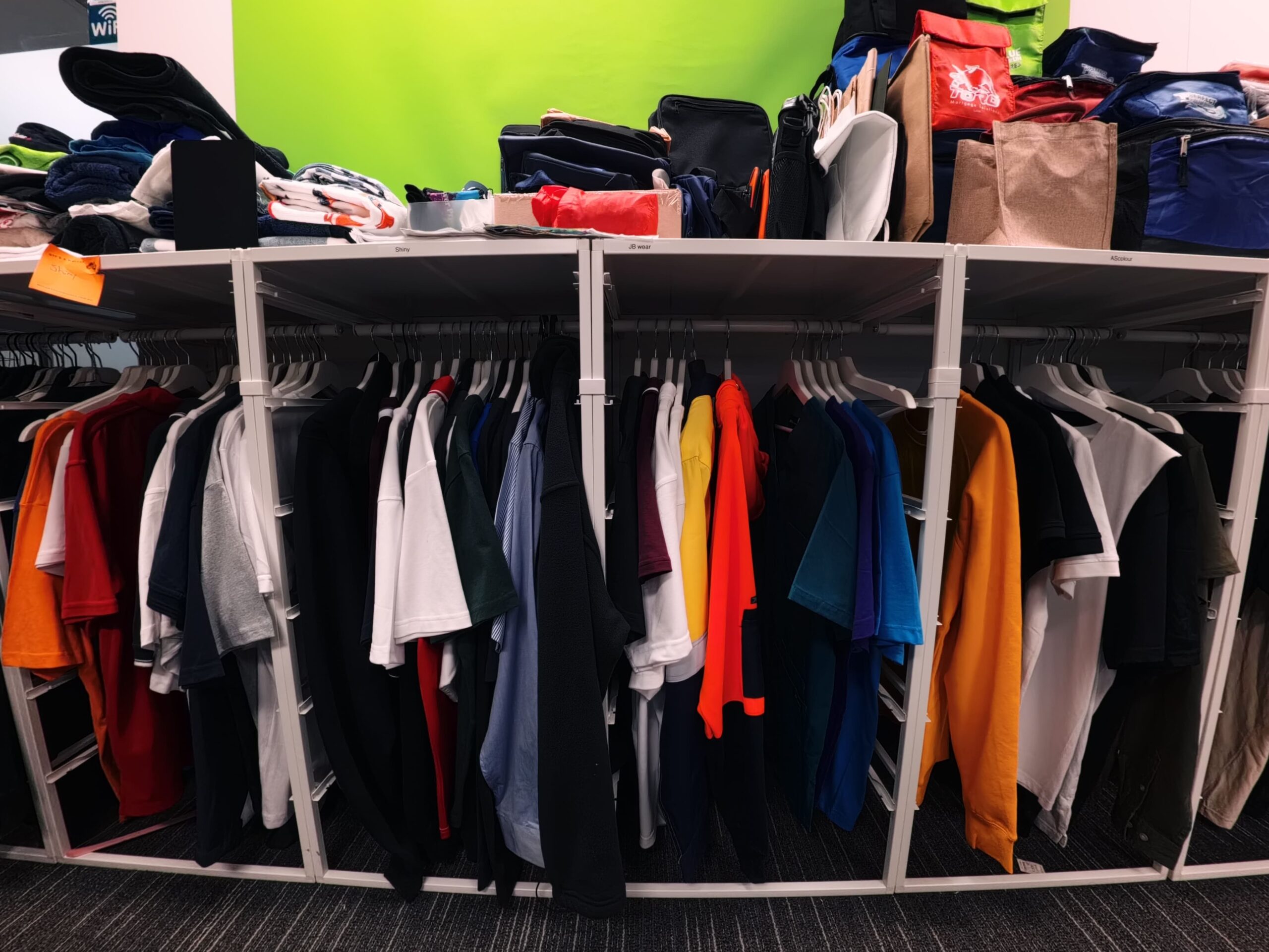 Assorted t-shirts in a showroom, with bags and folded clothes stacked neatly on the shelf above.