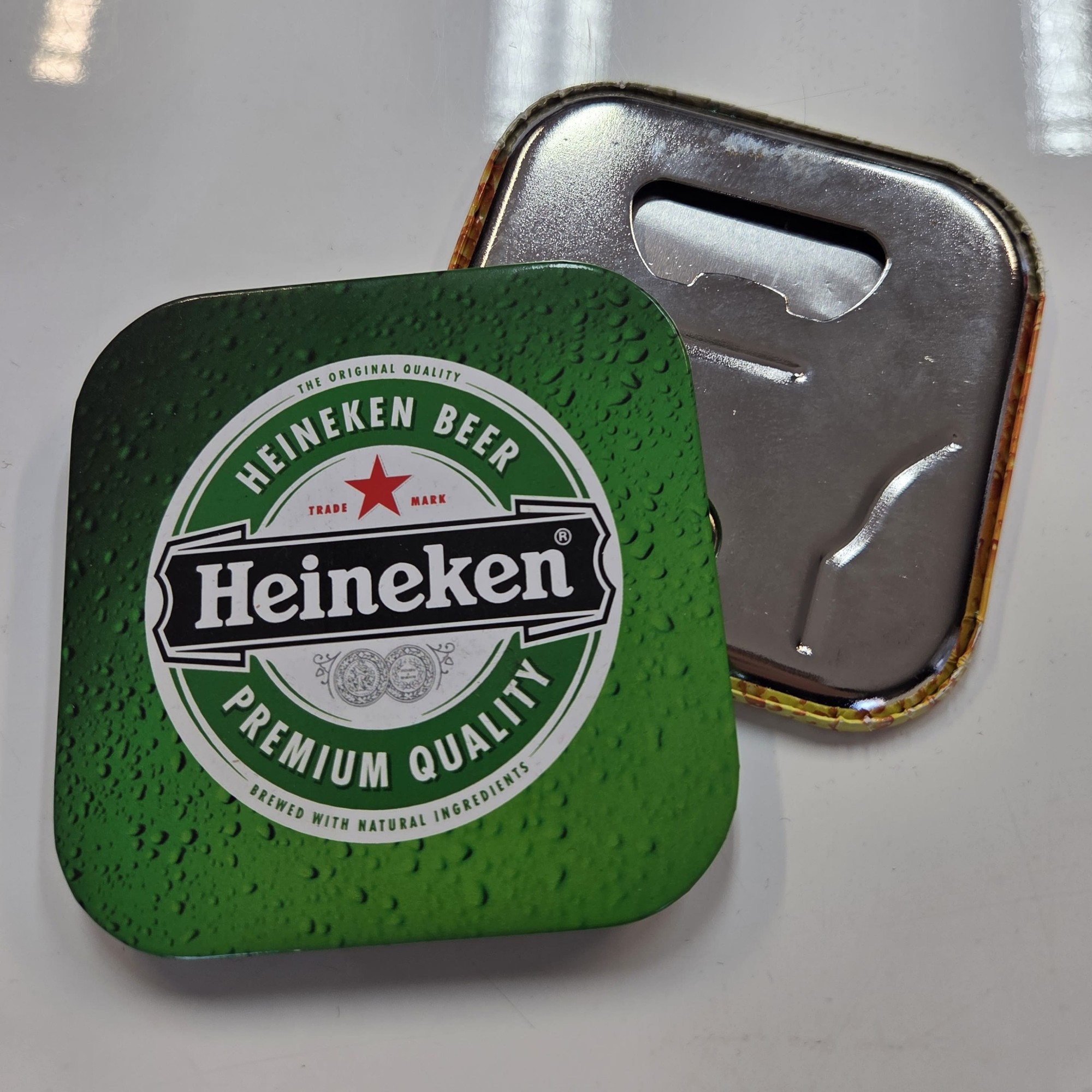 A Heineken-branded bottle opener featuring bold branding on a green front and metal back.