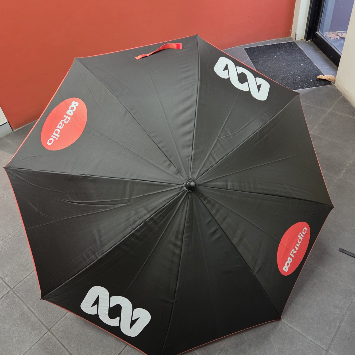 Black umbrella with ABC Radio logos highlights branding on a tiled floor by the red wall.