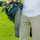 Person in khaki pants holding a golf bag with clubs on a grassy field.