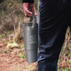 Person holding a gray ANKER travel mug with a handle while standing outdoors on a trail.