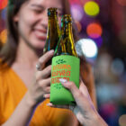 Two people clinking beer bottles with Autumn Harvest Festival sleeve, colorful lights behind.