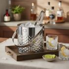 Stainless steel ice bucket with tongs, glasses, and lime slices on a wooden tray.