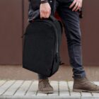 Person holding a black backpack, wearing dark jeans and brown boots, standing on a sidewalk.