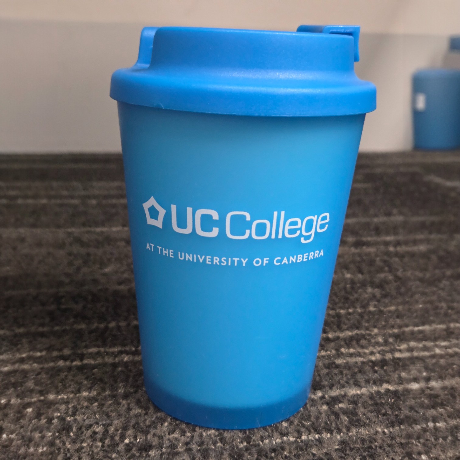 A blue UC College reusable cup showcasing print colour accuracy on a grey carpeted floor.