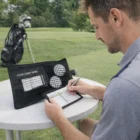 Man filling out scorecard with Corporate Golf Scorecard Holder, golf bag and course behind him.