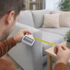 Person uses a House Tape Measure Keychain to measure a sofa by a wooden table in the living room.
