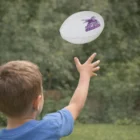 Child in blue shirt throws a Quanco Mini Rugby Ball outdoors with trees behind.