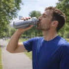 Man in blue shirt drinks from a Hydrate Water Bottle in a park on a sunny day.
