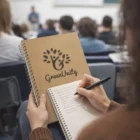 Student taking notes in class, holding a Brandable Eco A5 Note Pad with a tree logo on the cover.
