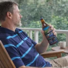 Man in a blue striped shirt holding a beer with Chugger Stubby Holders on a porch by the trees.