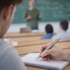 A student takes notes with Bronze Ball Pens while the teacher speaks at the blackboard.