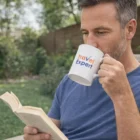 A man in blue reads a book outdoors, sipping from a Fine Bone China Mug labeled Travel Expert.