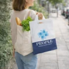 Woman carrying grocery bags and a Red Blue Black Tote Bag with vegetables, walking on sidewalk.