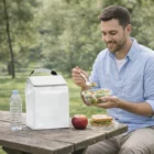Man eating salad at a picnic table with a Nomad Solo Cooler Bag, sandwich, apple, and water.
