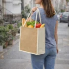 Woman walks city street with a Laminated Jute Bag Coloured filled with bread, lettuce, and vegetables.