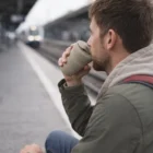Man at a train station sips from a Promo 350Ml Express Coffee Cup as the train approaches.