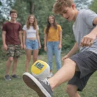 Teen kicks Colourful Branded Hacky Sacks as friends watch outdoors on grass.
