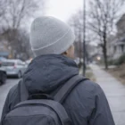 Person wearing a gray Dixie Beanies beanie and jacket with backpack walks on a cloudy sidewalk.